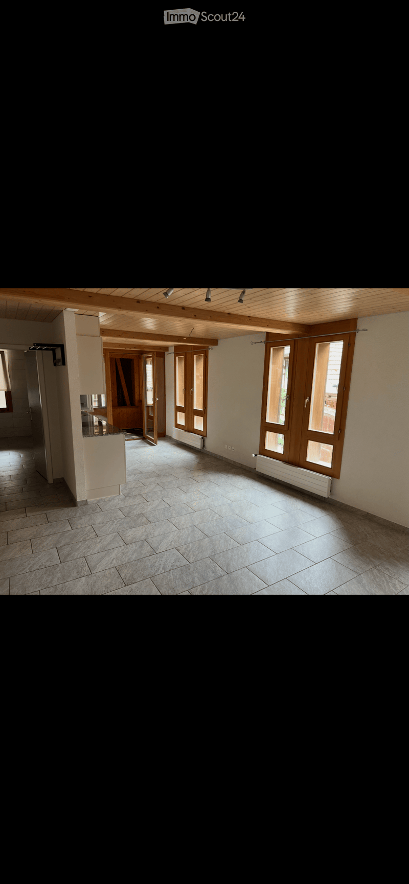 Empty room with tiled floor, wooden ceiling, large windows, and open doors leading to another area.