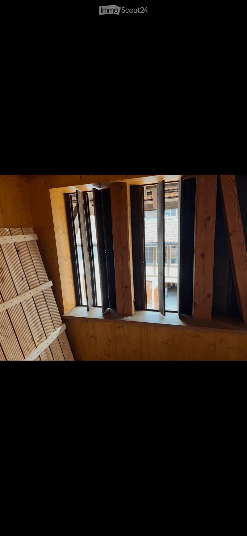 Empty room with wooden walls and a large window with shutters, showing a view of the outside area.
