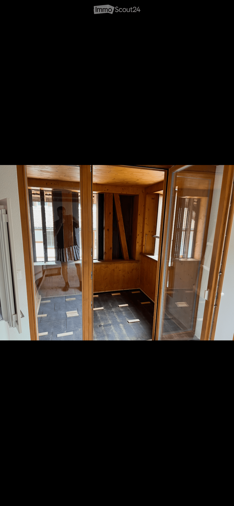 Empty room with wooden beams and a glass door leading to another room. The floor is unfinished, and the walls are bare.