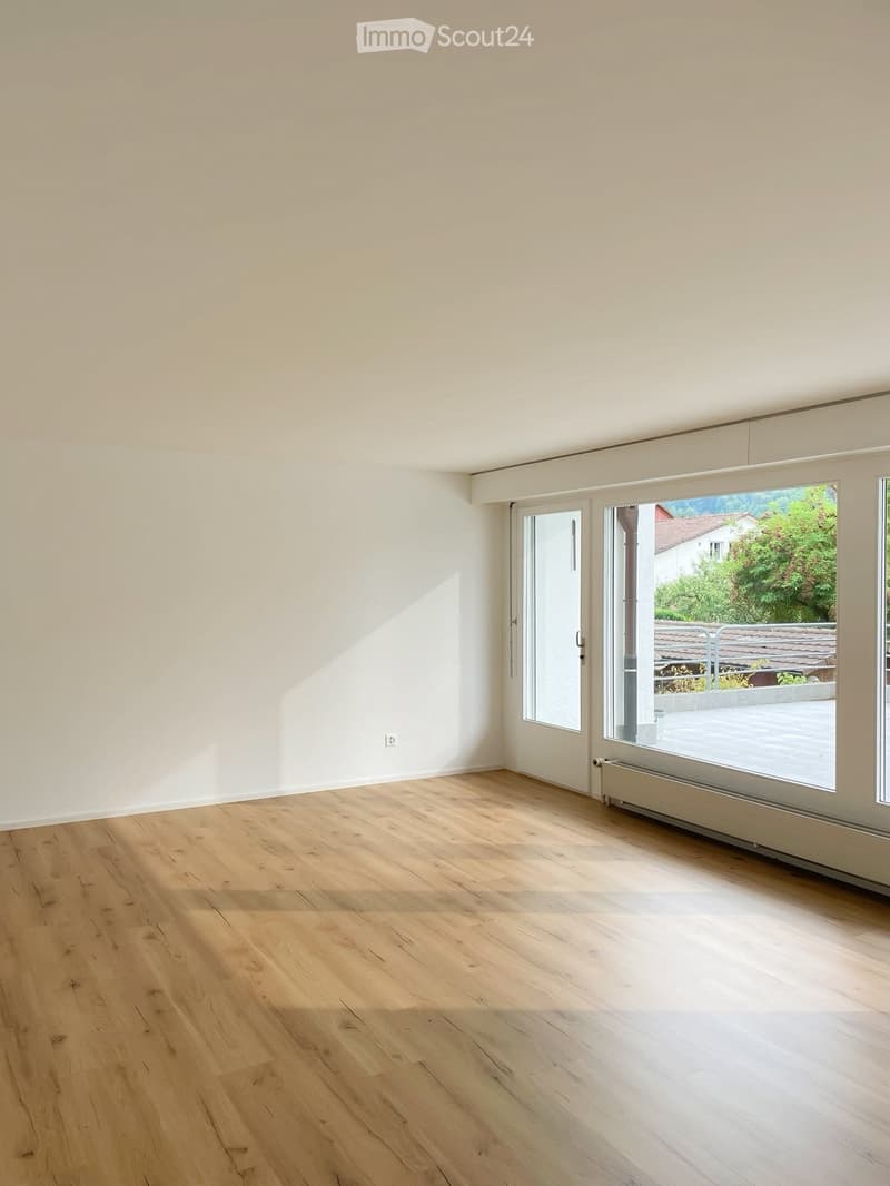 Empty room with light wooden flooring and white walls, featuring large windows and a sliding glass door leading to an outdoor area.