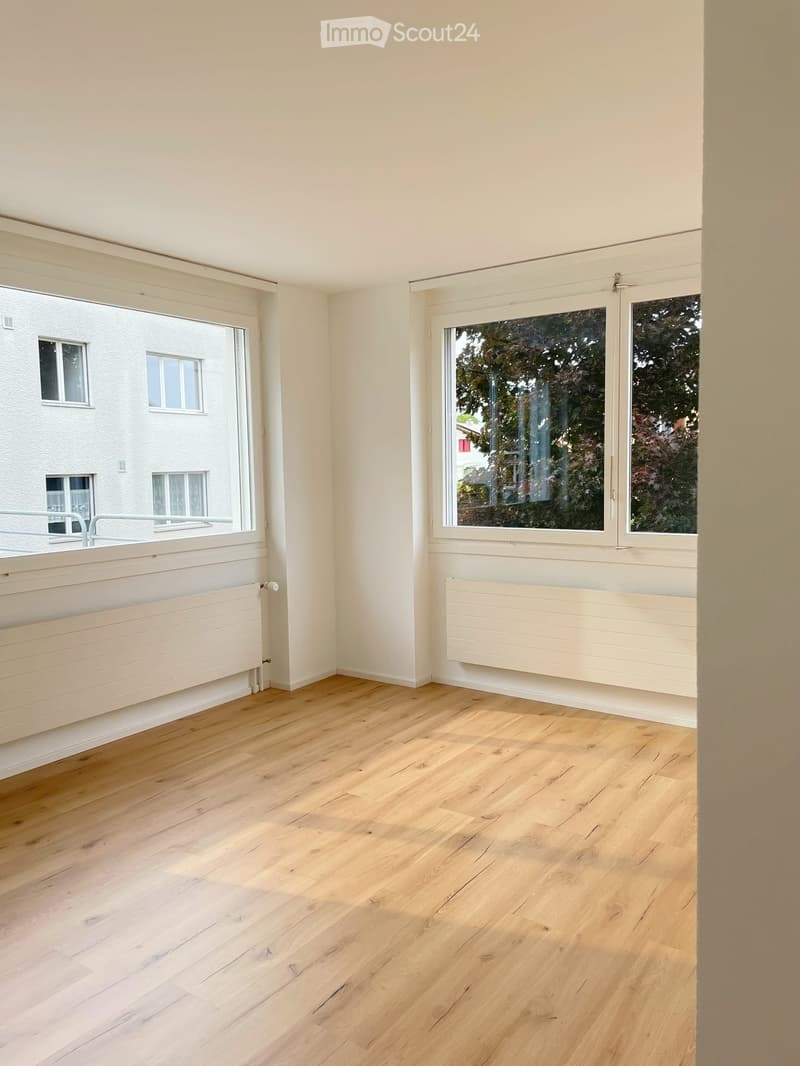 Empty room with light wooden flooring, white walls, large windows, and radiators. No furniture or equipment present.