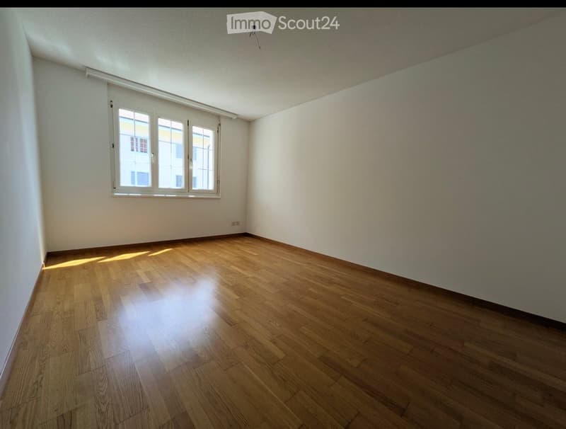 Empty room with wooden flooring and white walls, featuring a large window with blinds and natural light streaming in.