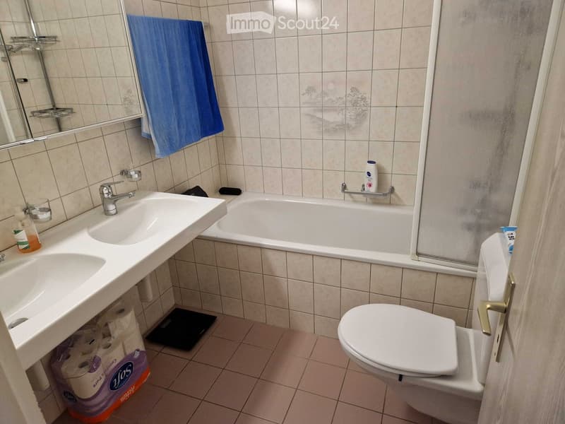 Bathroom with a double sink, bathtub, toilet, mirror, and a blue towel hanging on the wall. Toilet paper and a bottle of lotion are visible on the floor and bathtub ledge.