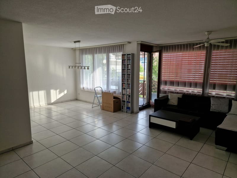 Bright living room with tiled floor, large windows, a black sofa, coffee table, bookcase, and a folding chair.