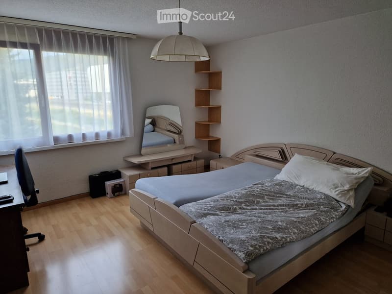 A well-lit bedroom with a wooden bed, nightstand, mirror, shelves, and a hanging lamp. The room features a large window with sheer curtains and a wooden floor.