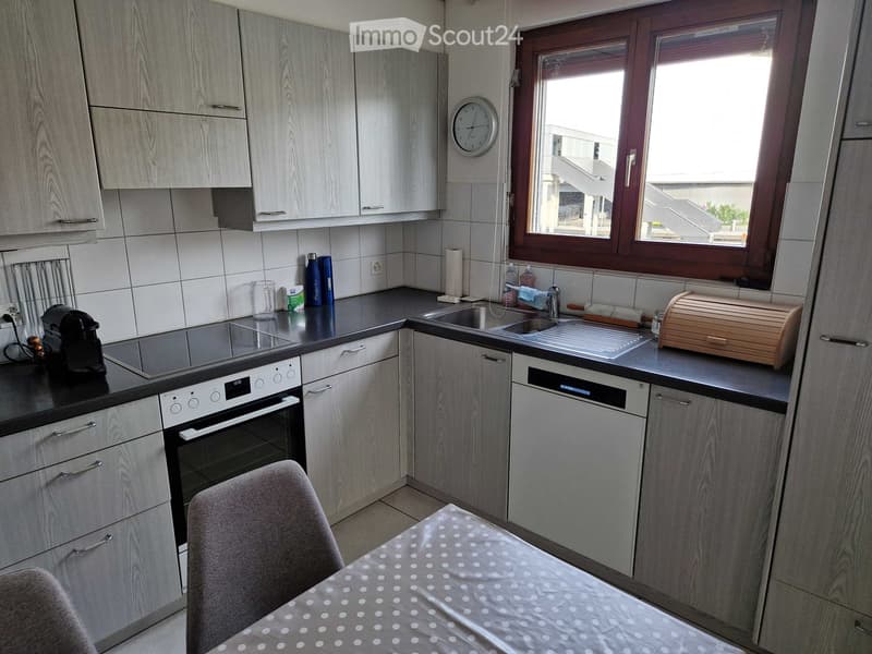 Modern kitchen with light wood cabinets, stainless steel appliances, and a black countertop. Features an oven, dishwasher, sink, and a small dining area with a table and chair.