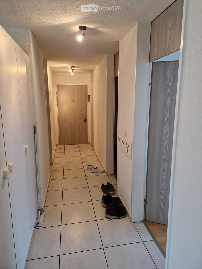 A narrow hallway with tiled flooring, white walls, and several doors. Shoes are placed on the floor, and a broom leans against the wall.
