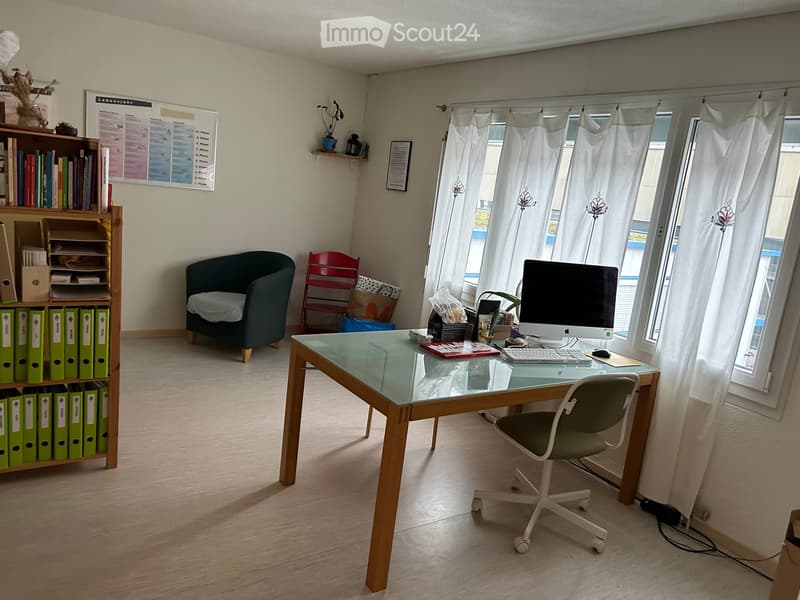 Well-lit office space with a glass desk, office chair, computer setup, a small sofa, a red chair, and a bookshelf filled with books and filing cabinets.