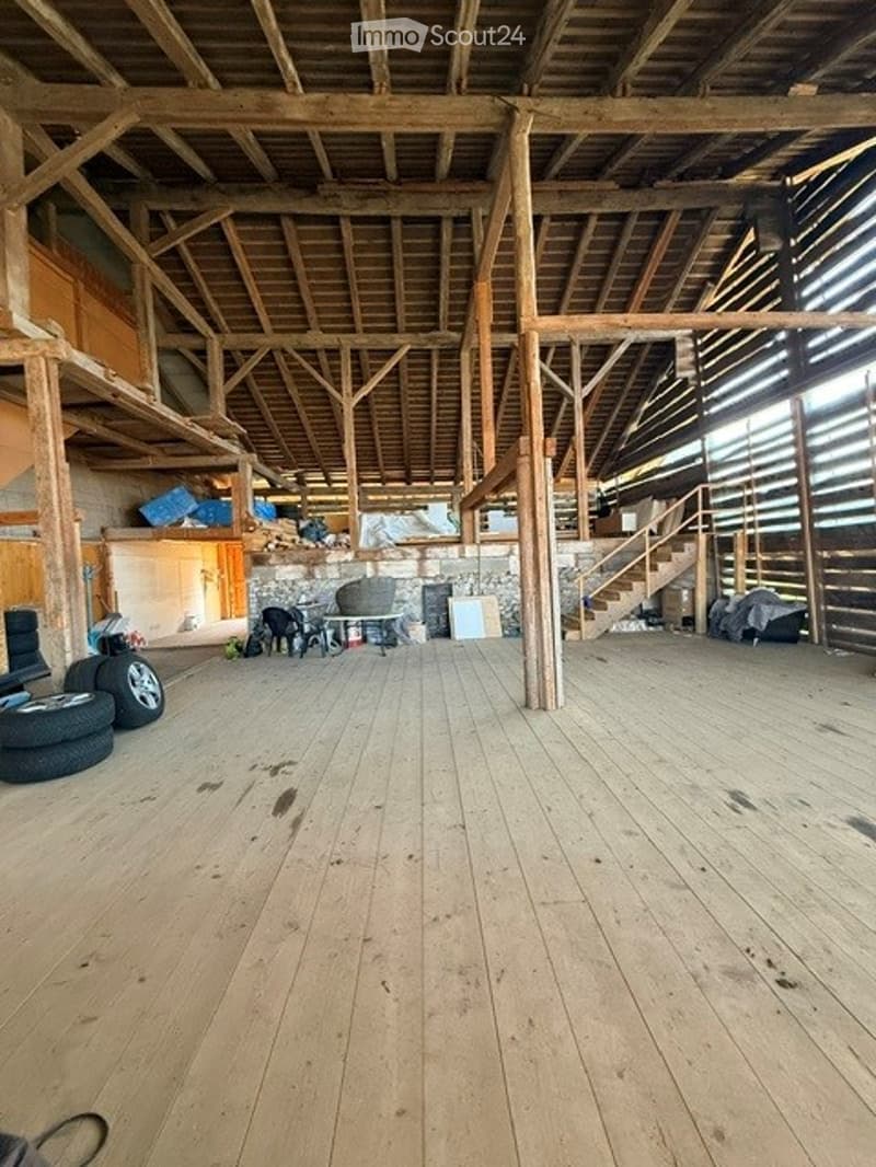 Large, open, wooden-floored space with exposed wooden beams and pillars. Minimal furnishings, including a few chairs and a small table, with storage items and tires visible.