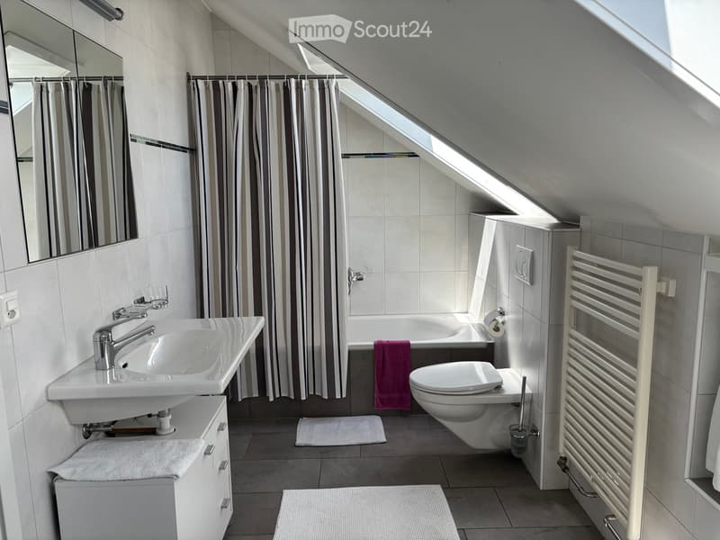 Modern bathroom with a sink, mirror, bathtub, toilet, radiator, and towels. Features a skylight window and striped curtains.