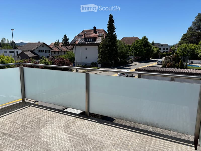 Balcony with glass railing, tiled floor, overlooking a residential street with houses, trees, and parked cars.