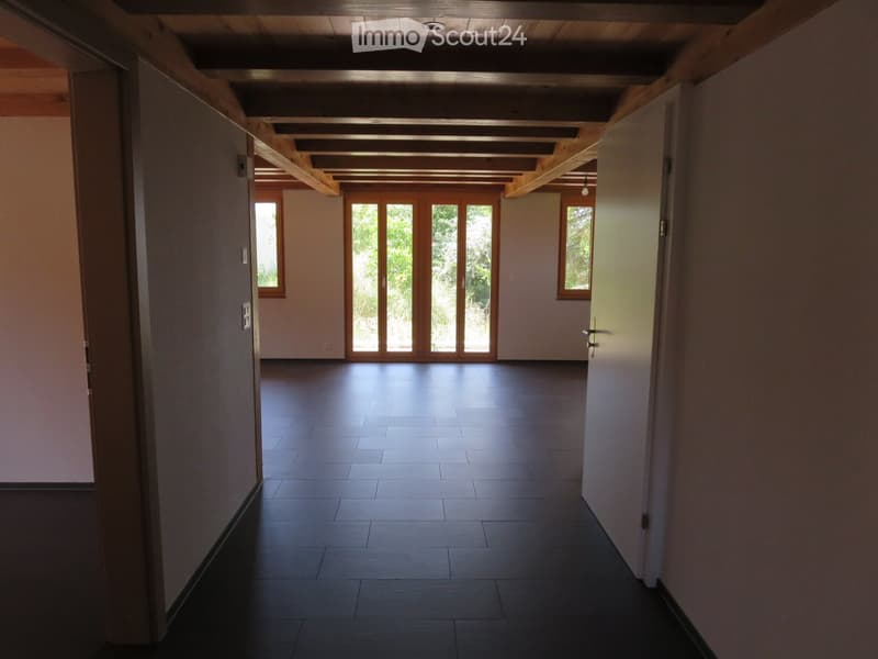 Empty hallway with wooden ceiling beams, dark tiled floor, and large glass doors leading outside.