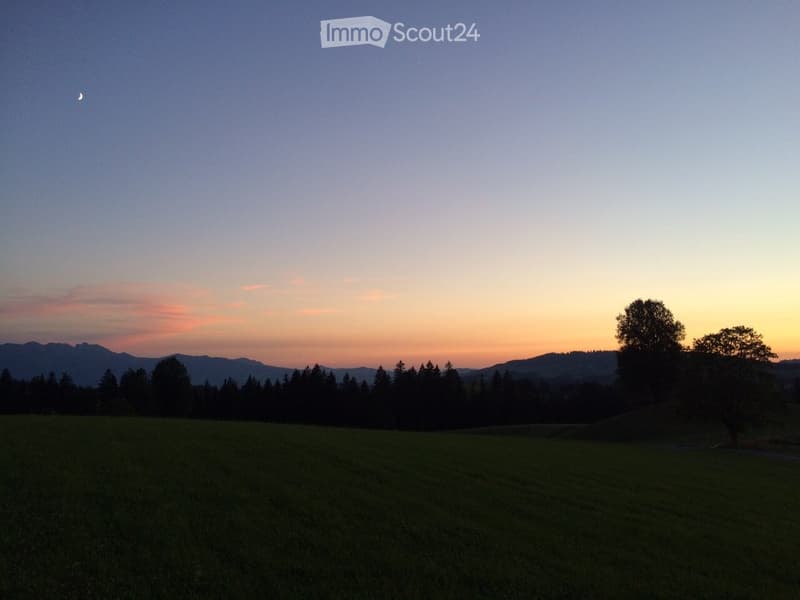 A serene landscape at sunset with a grassy field, scattered trees, and distant mountains under a twilight sky.