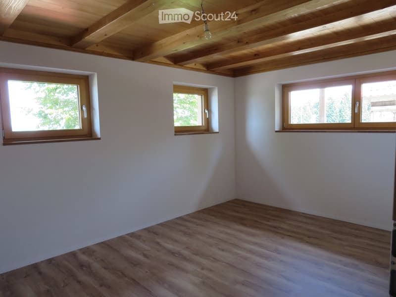 Empty room with light wooden flooring, white walls, and three windows with wooden frames. The ceiling features exposed wooden beams.