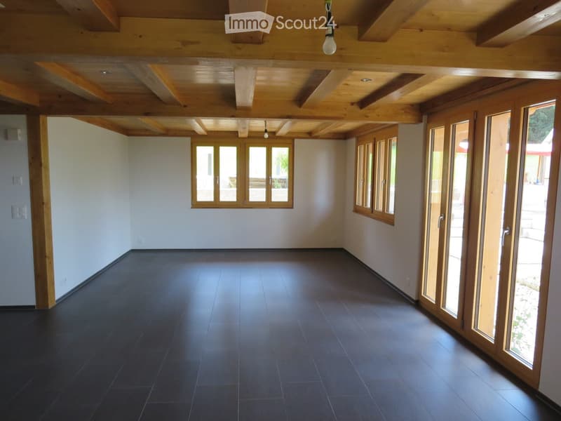 Empty room with dark tiled floor, light-colored walls, wooden ceiling beams, and large windows with wooden frames. No furniture or equipment present.