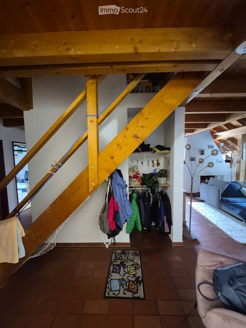 A cozy living space with exposed wooden beams, a staircase leading to an upper level, a clothing rack with jackets and bags, a sofa, a chair, and a floor lamp.