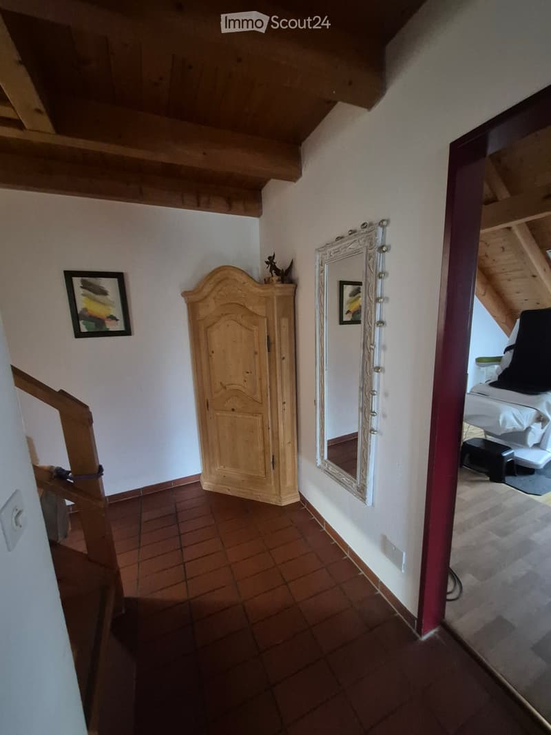 A narrow hallway with exposed wooden beams, a large ornate mirror, a wooden cabinet, and a framed picture on the wall. The hallway leads to a bedroom with a bed and nightstand visible in the background.