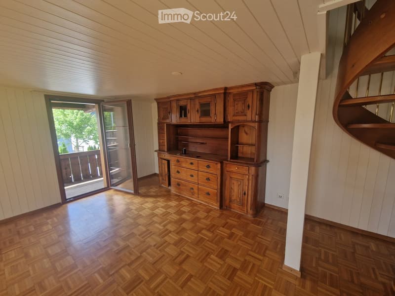 Spacious room with wooden parquet flooring, large wooden cabinet, and a spiral staircase leading upwards. Features a large window with a view of the balcony.