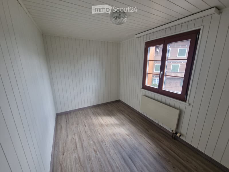 Empty room with wooden flooring, white paneled walls, a single window with brown frame, and a radiator below the window.