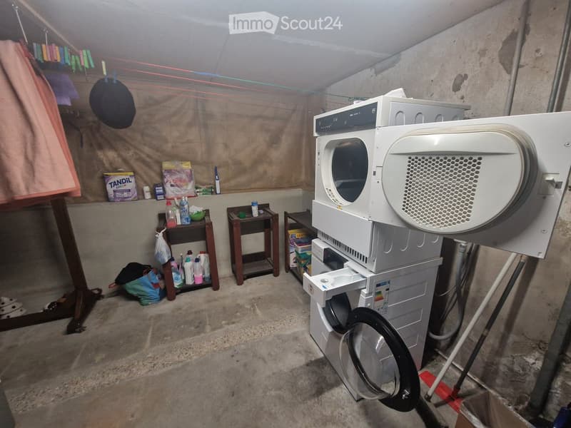 A small utility room with a washing machine, microwave, and various storage items including bottles and a hat on a shelf.