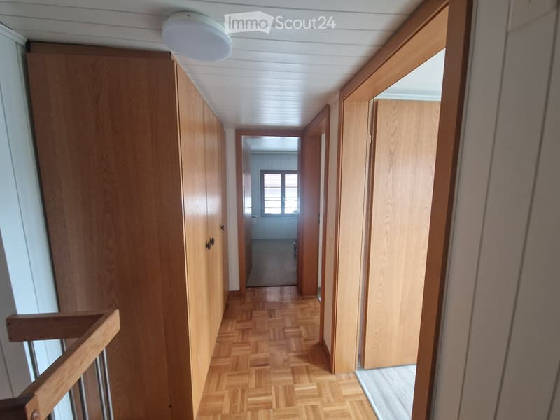 A narrow hallway with wooden doors, herringbone-patterned wooden flooring, and a white ceiling light fixture.