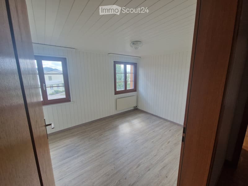 Empty room with wooden flooring, white paneled walls, two windows, and a radiator. The ceiling has a light fixture.