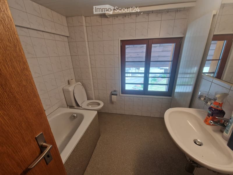Bathroom with tiled walls, a toilet, a bathtub, a sink, and a mirror. The room has a window with blinds and a wooden door partially visible.