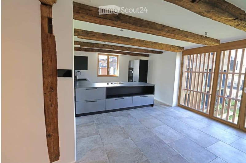 Modern kitchen with exposed wooden beams, grey cabinets, a central island, and large windows with wooden shutters.