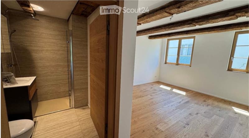 Empty room with wooden flooring, white walls, and exposed wooden beams on the ceiling. A separate bathroom with a sink, toilet, and glass shower enclosure is visible through an open doorway.