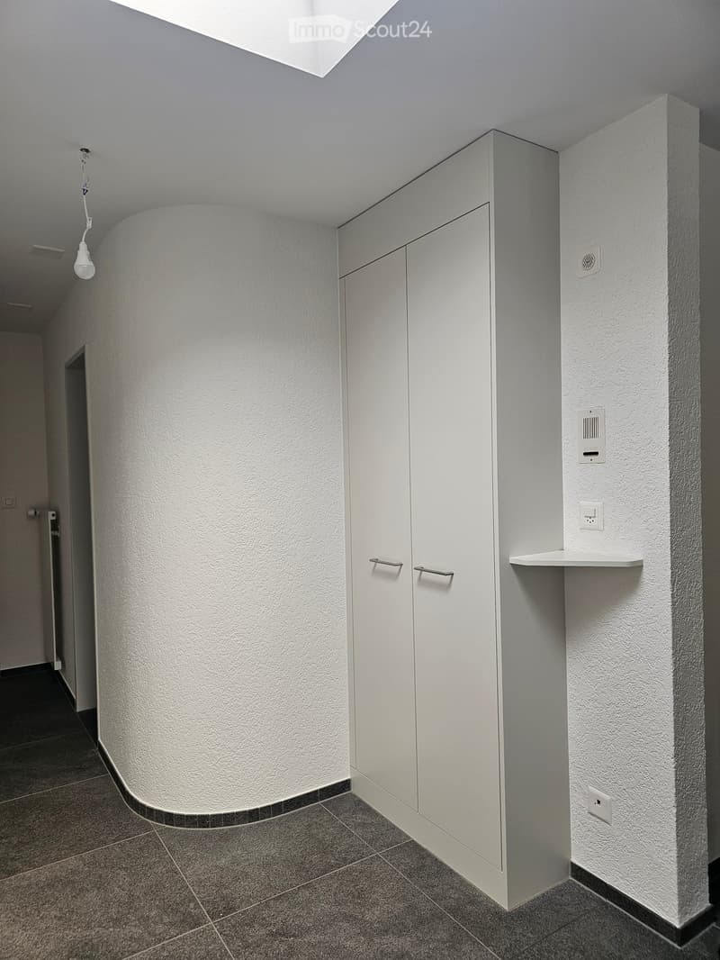 Clean, modern hallway with white walls, grey tiled floor, built-in white storage unit with double doors, and a small shelf on the wall.
