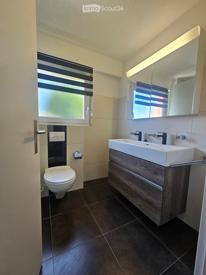 Modern bathroom with dark tiled floor, white toilet, white sink with dark wooden cabinet, mirror, and window with blinds.