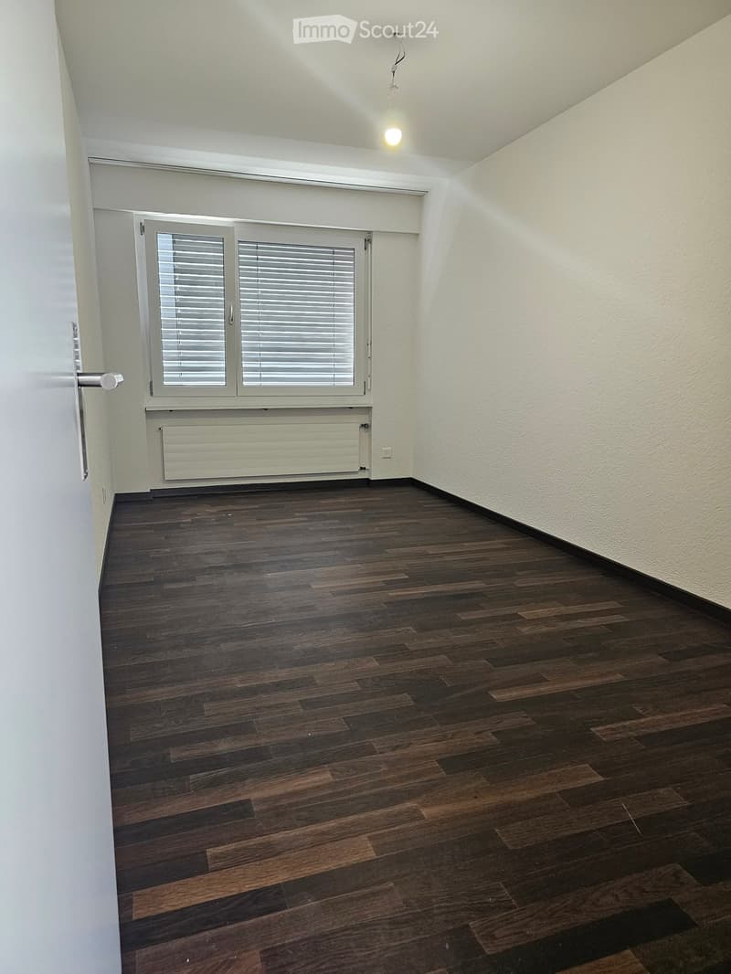 Empty room with wooden flooring, white walls, a window with blinds, and a radiator below it.