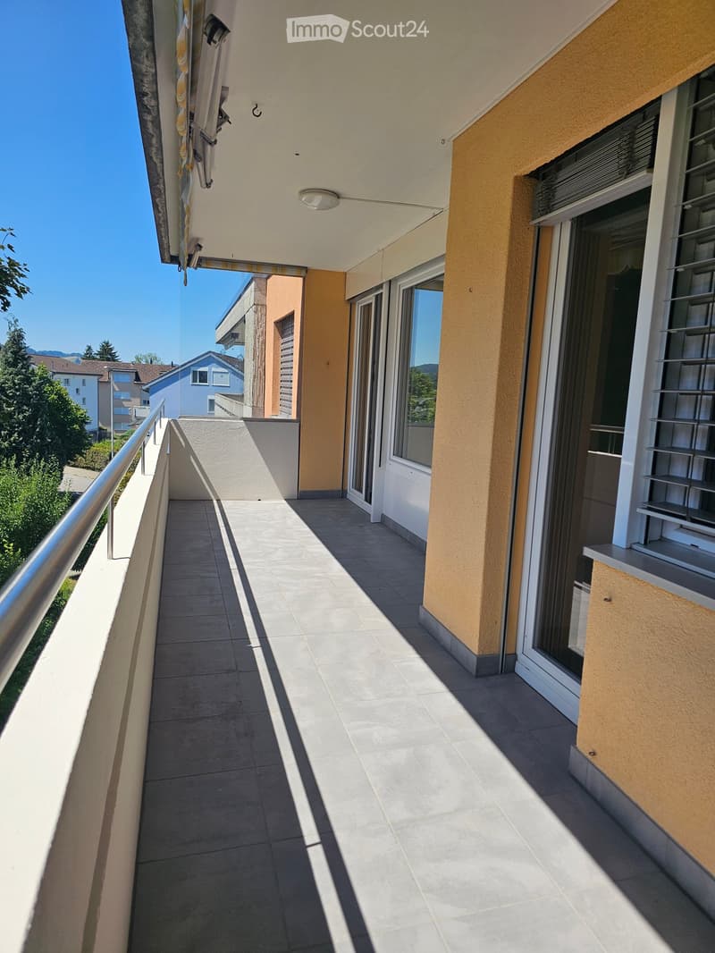 Balcony with tiled floor, metal railing, and large windows overlooking a residential area with trees and buildings.