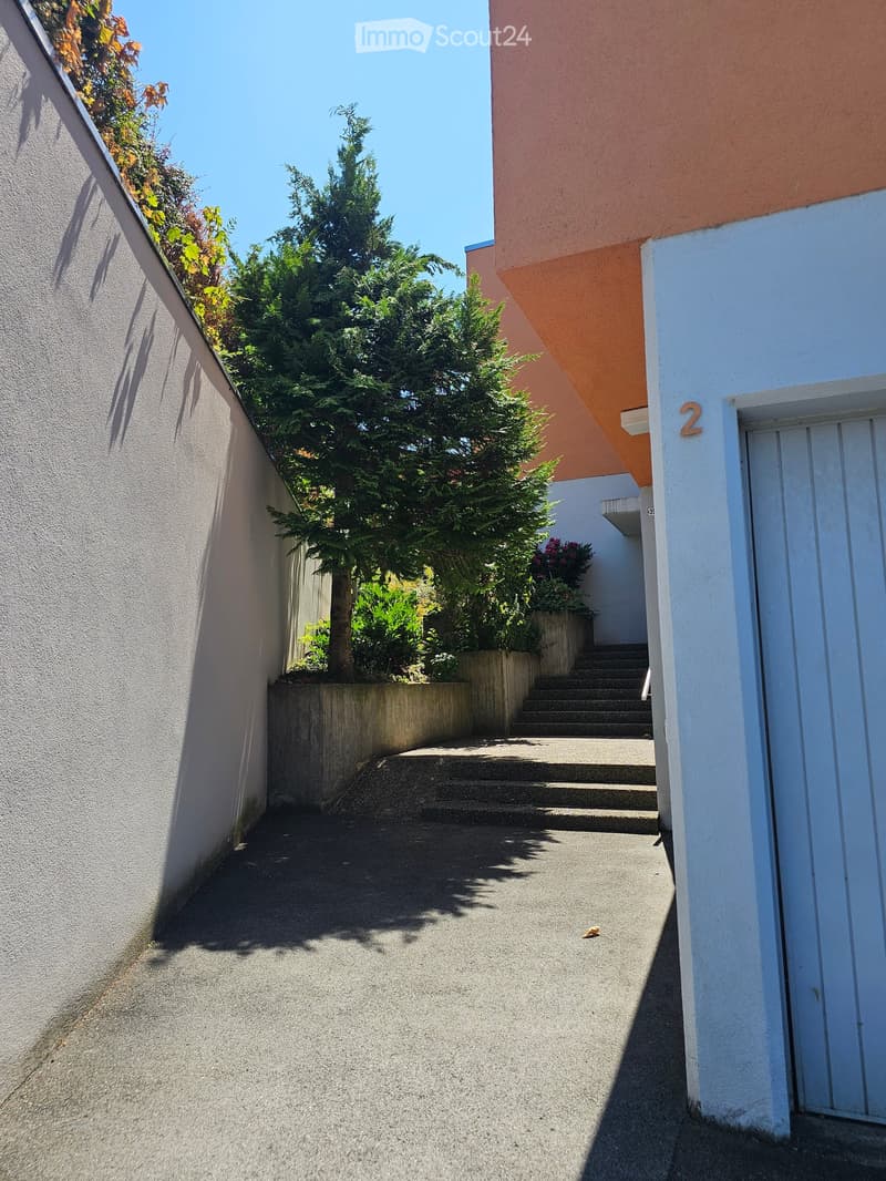 A narrow concrete pathway between two buildings, with steps leading up to a higher level and greenery visible in the background.