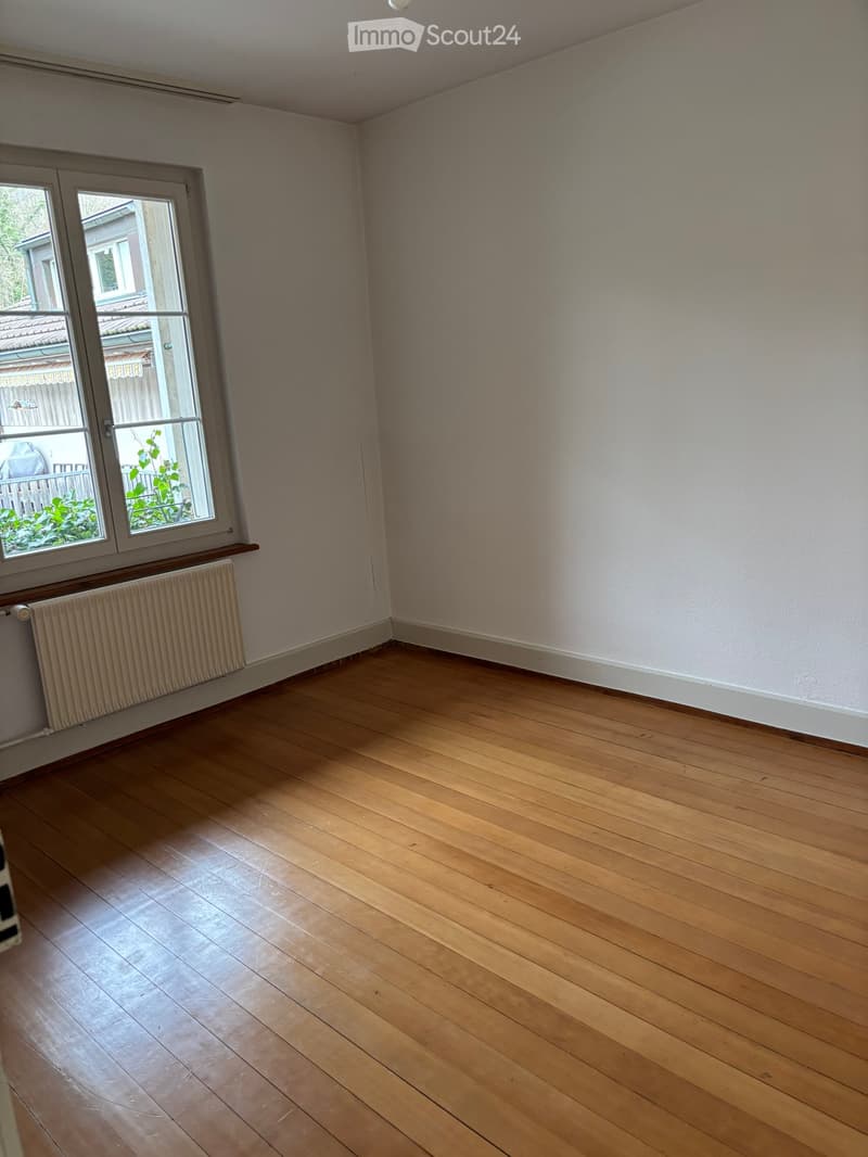 Empty room with wooden flooring, white walls, and a window with a radiator below it.