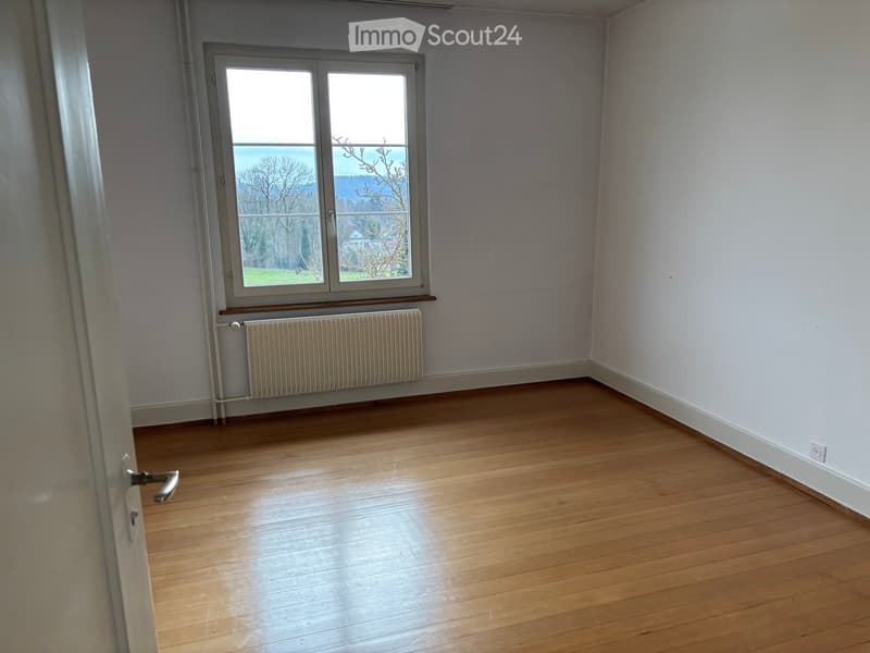 Empty room with wooden flooring, white walls, a window with a view, and a radiator below the window.