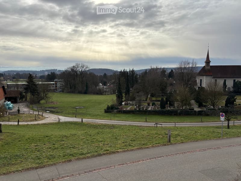 Rural landscape with a church, cemetery, grassy fields, trees, and a road.