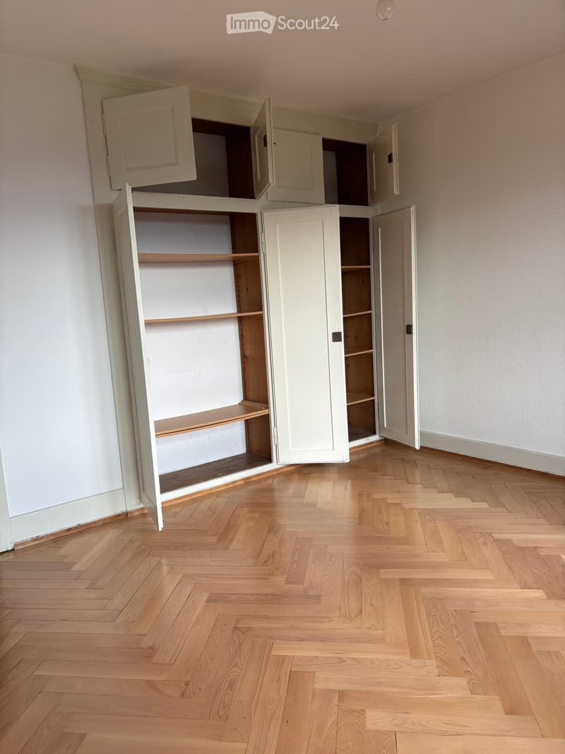 Empty room with light wooden herringbone flooring and white walls. Open built-in cabinetry with shelves is visible on one wall.