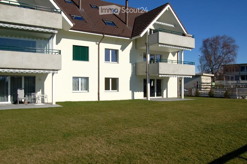 Two-story house with white walls, brown roof, multiple windows, and balconies. A small patio area with a table and chairs is visible on the left side.