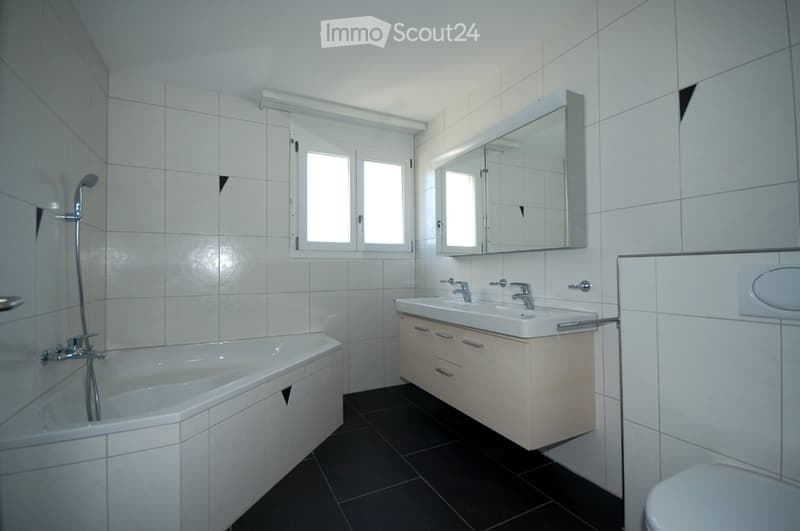 Modern bathroom with white tiles, a bathtub, a double sink with a mirrored cabinet, and a toilet. The room features a window with frosted glass panes and a sleek, contemporary design.