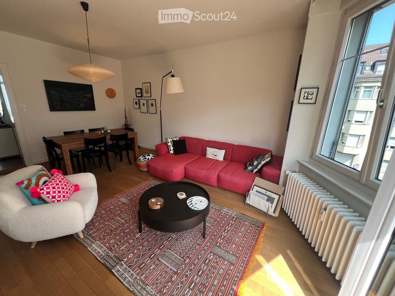 Modern living room with a red sectional sofa, a round coffee table, a grey armchair, a dining area with a wooden table and chairs, and a colorful rug.
