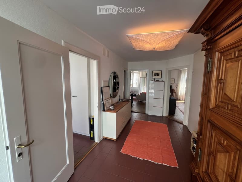A well-lit hallway with tiled flooring, a red runner, and several doors leading to different rooms. Visible features include a staircase on the right and a small console table with a mirror and decorative items on the left.