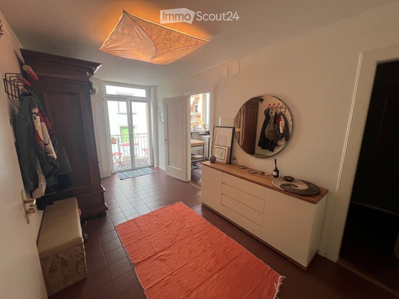 A furnished room with a bed, a mirror, a cabinet, a small table, a stool, a chair, and a coat rack. The room has tiled flooring and a slanted ceiling with a hanging light fixture.
