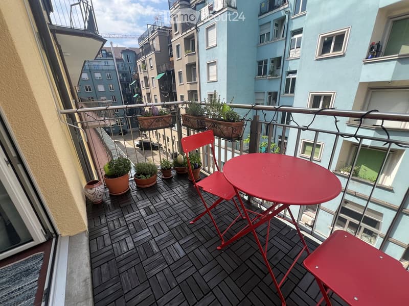 Balcony with wooden decking, red metal table and chairs, several potted plants, and a view of the city street.