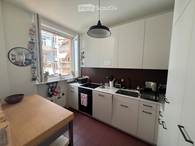 Modern kitchen with white cabinets, a wooden table, a sink, an oven, and a dishwasher. The space features a large window with a view of the street and several potted plants on the windowsill.