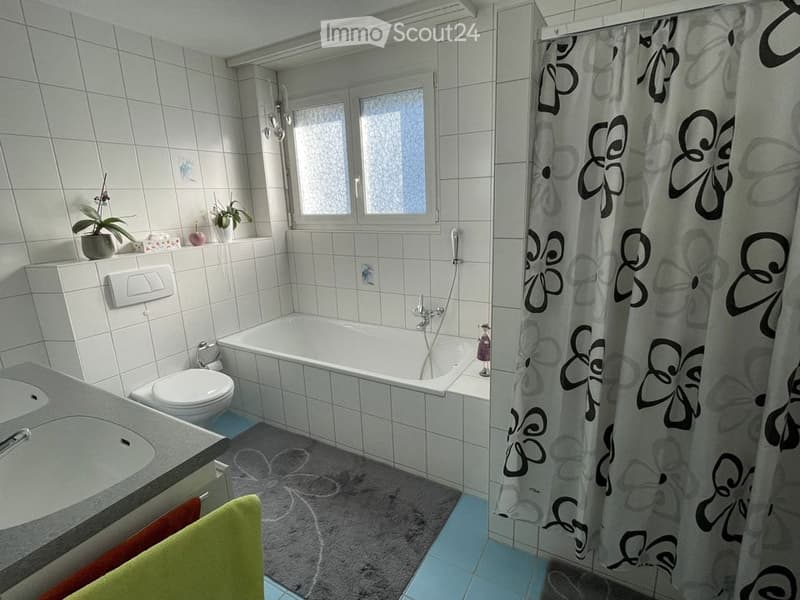 Bathroom with a bathtub, toilet, sink, and a shower curtain. The room features tiled walls and a window above the bathtub. There are small plants and towels visible.