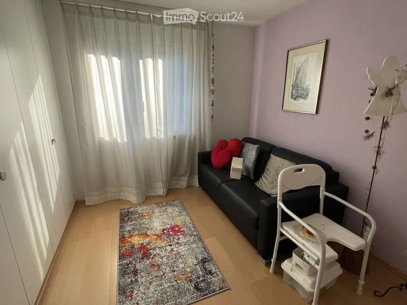 A cozy living room with a black sofa, colorful cushions, a white chair, a colorful rug, and a floor lamp. The room has light purple walls, large windows with white curtains, and a framed picture on the wall.