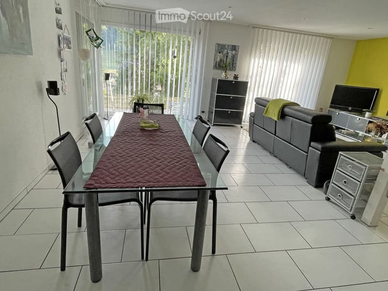 Modern dining and living area with a glass dining table, six chairs, a dark grey sofa, a TV stand, a small cabinet, and a floor lamp. The space features large windows with vertical blinds and a bright yellow accent wall.