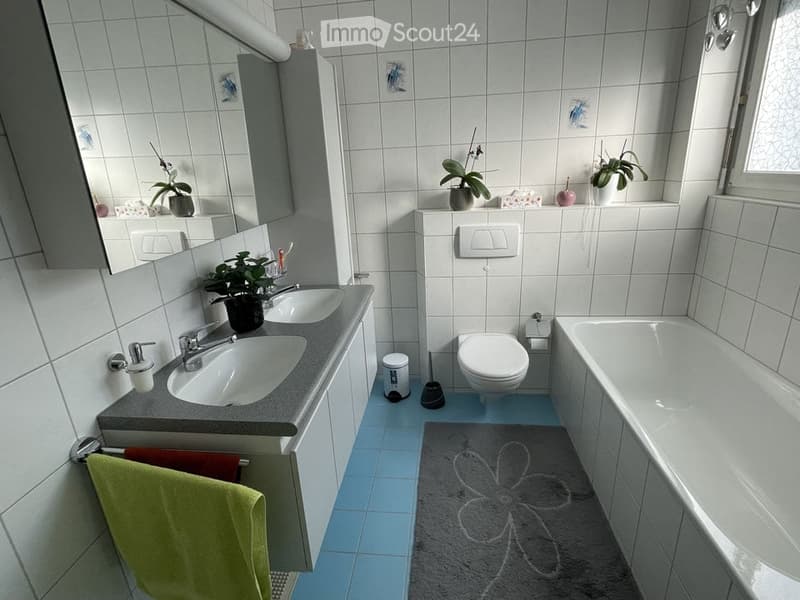 A modern bathroom with white tiled walls and blue tiled floor, featuring a double sink vanity, a toilet, a bathtub, a large mirror, and decorative plants on the windowsill.