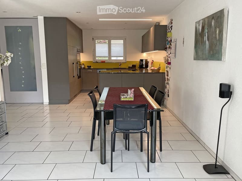 Modern dining area with a glass table and black chairs, adjacent to a sleek kitchen with grey cabinets, a yellow accent wall, and stainless steel appliances.